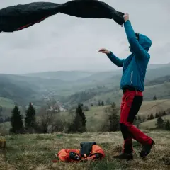 Osoba w niebieskiej kurtce i czerwonych spodniach rozkłada śpiwór puchowy czy syntetyczny na tle górskiego krajobrazu.
