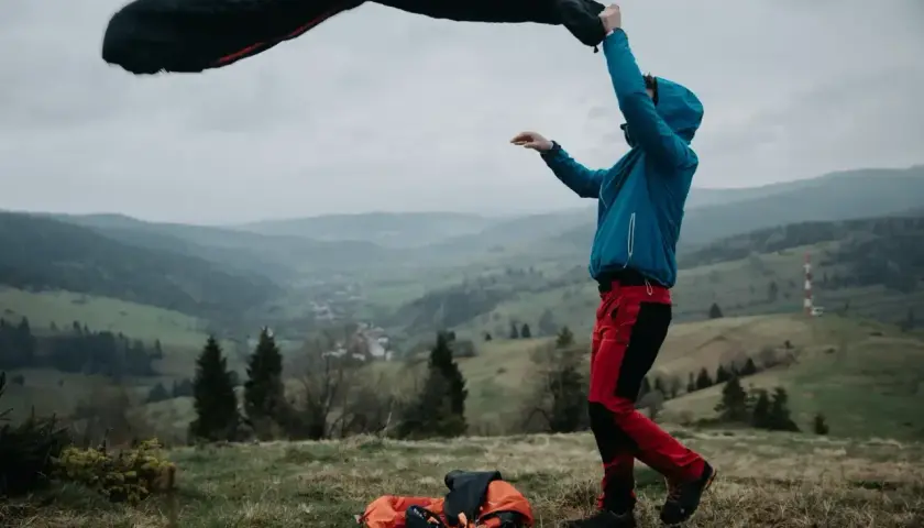 Osoba w niebieskiej kurtce i czerwonych spodniach rozkłada śpiwór turystyczny na tle górskiego krajobrazu.