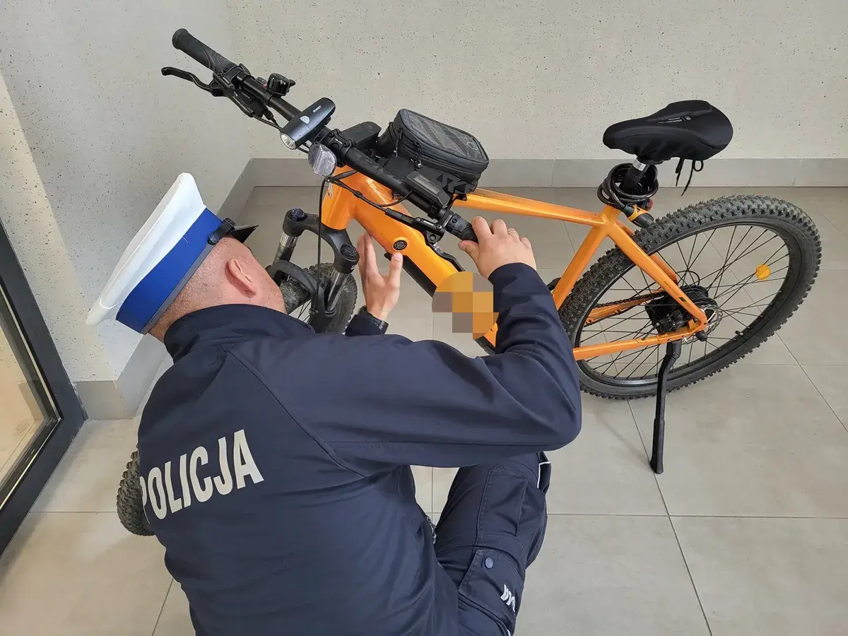 Policjant w czapce bada pomarańczowy rower elektryczny, próbując zrozumieć, jak działa rower elektryczny.