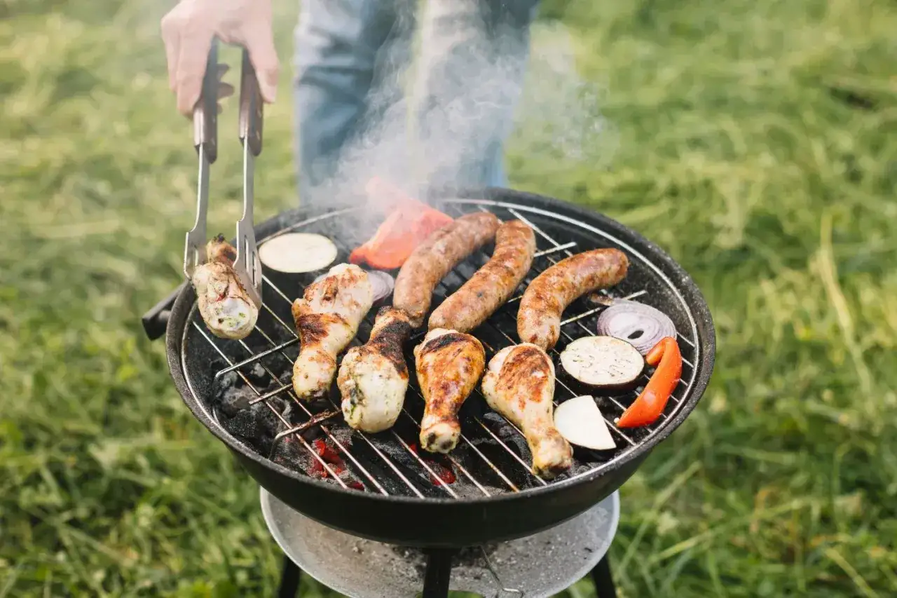 Ręka z szczypcami obraca kurczaka na grillu. Kiełbaski, papryka i cukinia też się pieką. Idealny sposób, jak rozpalić grilla na letni wieczór.