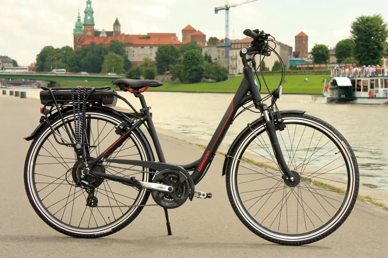 Czarny rower z bagażnikiem i baterią, gotowy do przeróbki na elektryczny. W tle Wawel i Wisła.
