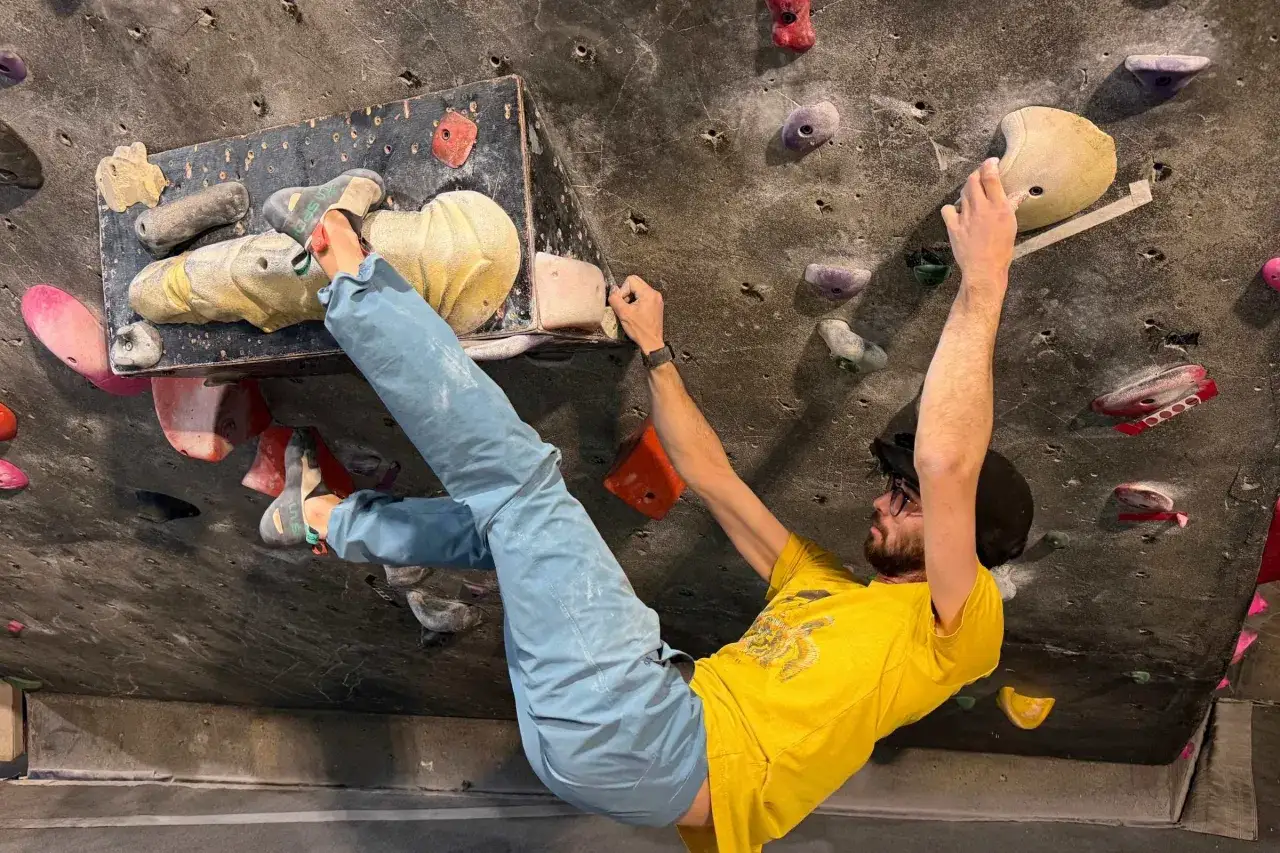 Mężczyzna w ż&oacute;łtej koszulce wspina się po ścianie z chwytami. To właśnie bouldering &ndash; wspinaczka bez liny na niskich ścianach.