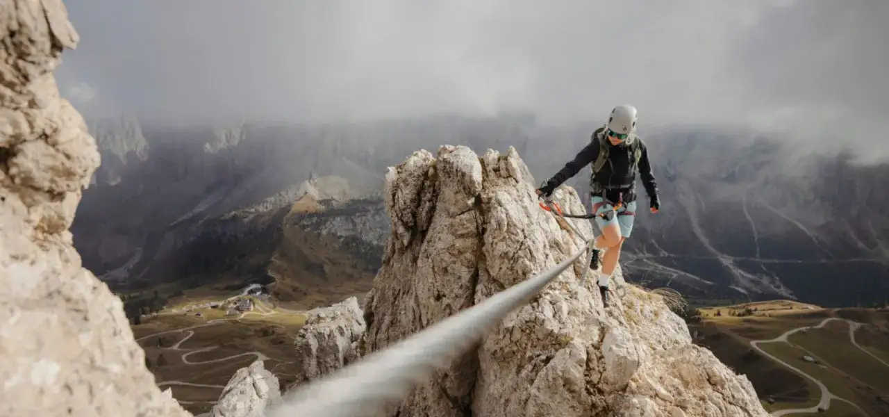Kobieta w kasku i sprzęcie wspinaczkowym pokonuje via ferrata dla początkujących. Widok na górzysty krajobraz.