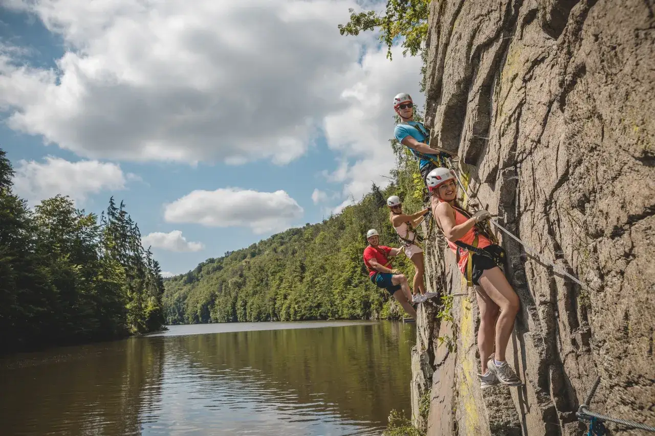 Grupa przyjaci&oacute;ł pokonuje via ferrata w Czechach, wspinając się po skalnej ścianie nad malowniczym jeziorem.